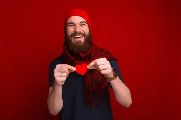 Młody brodaty mężczyzna trzymający serce z papieru i uśmiechnięty przy czerwonej ścianie. — Zdjęcie stockowe