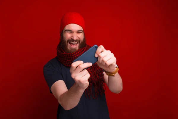 Radosny brodaty mężczyzna gra w swoim smartfonie w pobliżu czerwonego tła. — Zdjęcie stockowe