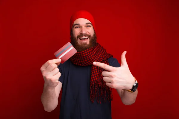 Bearded man is smiling, looking at the camera and pointing to credit card near red wall.