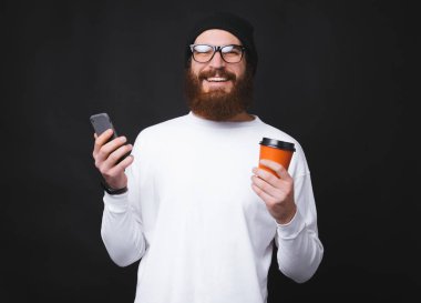 Sakallı hippi adam kameraya bakıp gülümserken elinde bir fincan kahve ve akıllı telefon tutuyor.