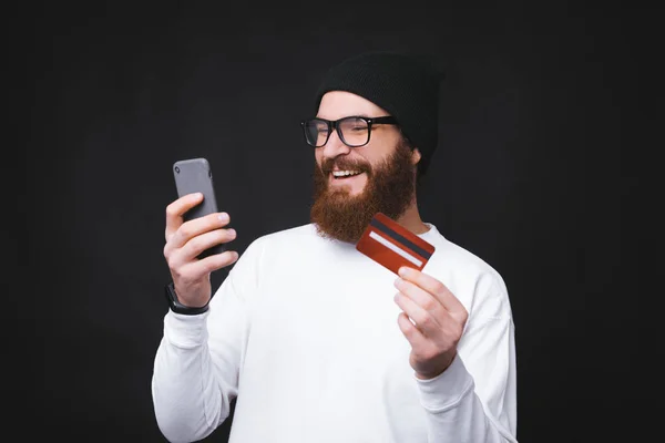 Joven hombre guapo con barba haciendo compras en línea con teléfono y tarjeta de crédito — Foto de Stock