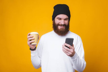 Telefonunu kullanan ve elinde kahve tutan neşeli bir gencin fotoğrafı.