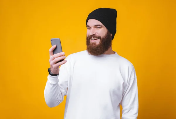 Ung glad trendig man med skägg med sin telefon över gul bakgrund — Stockfoto