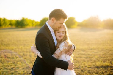 Birbirine aşık genç bir çift gün batımında tarlada sarılıp gülümsüyor..