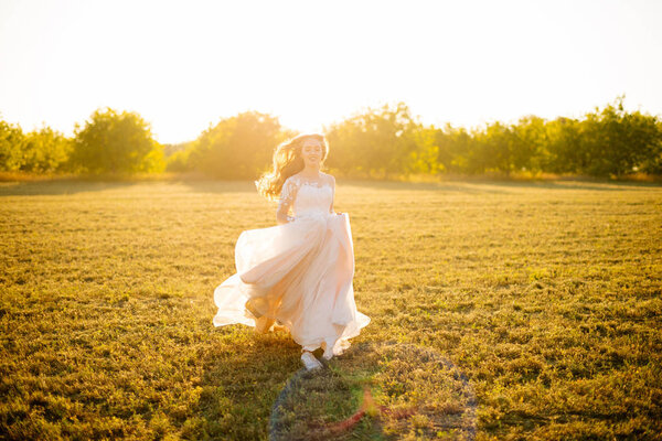 Young happy bride is running in front of the camera on the field at sunset.