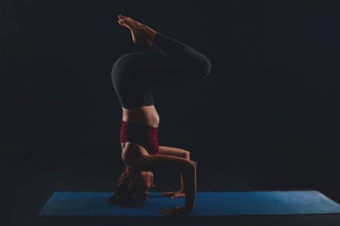 Stüdyoda yoga yapan genç bir kadın. Arka planda üç ayaklı amuda kalkıyor.