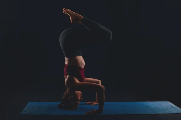 Mujer joven practicando yoga en el estudio sobre fondo oscuro trípode cabecera —  Fotos de Stock