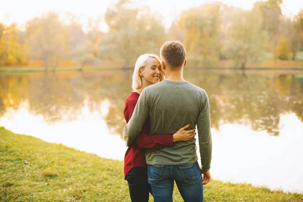 Foto de pareja joven y feliz abrazándose al atardecer cerca del lago durante —  Fotos de Stock