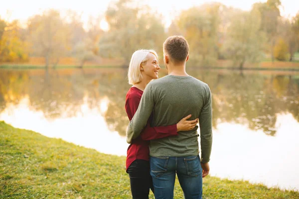 Foto de pareja joven y feliz abrazándose al atardecer cerca del lago durante — Foto de Stock