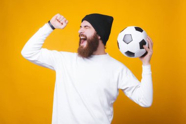 Sakallı neşeli adam bağırıyor ve sarı duvarda bir futbol topu tutuyor..