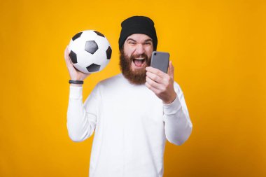 Genç sakallı adam selfie çekiyor ve sarı duvarın yanında bir futbol topu tutuyor..