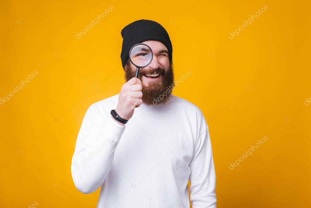 Young bearded man is looking through a magnifying glass and smiling near yellow wall.