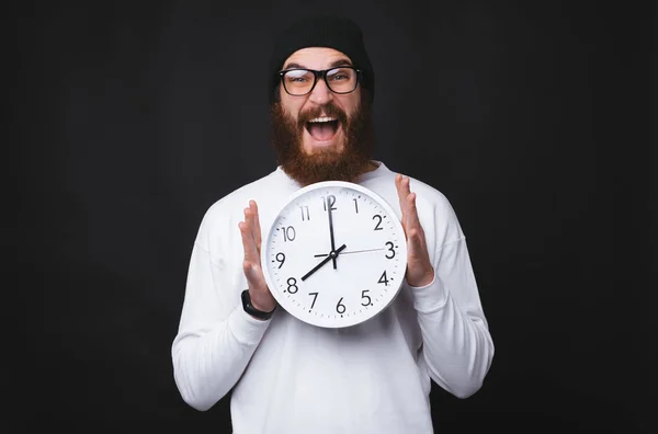 Un hombre joven y barbudo excitado sostiene un gran reloj blanco sobre fondo negro . — Foto de Stock