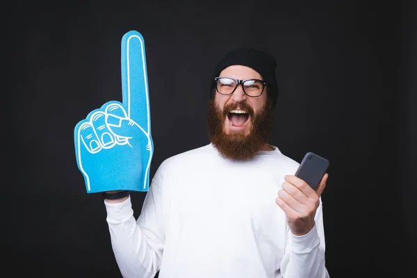 Un bărbat frumos cu barbă ține emoționat o mănușă de ventilator și un smartphone . — Fotografie, imagine de stoc