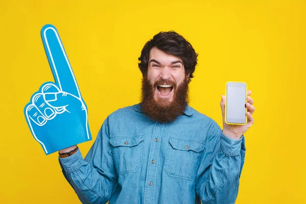 Hombre sorprendido gritando objetivo apuntando con el guante del ventilador y mostrando la pantalla del teléfono inteligente —  Fotos de Stock