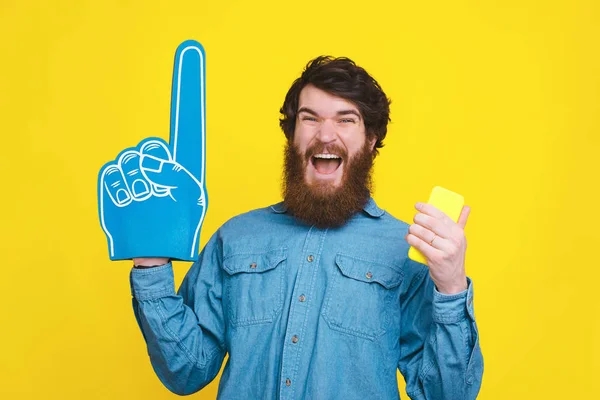 Hombre sorprendido en camisa azul celebrando la victoria y apuntando hacia arriba con el guante de espuma del ventilador y sosteniendo el teléfono inteligente —  Fotos de Stock