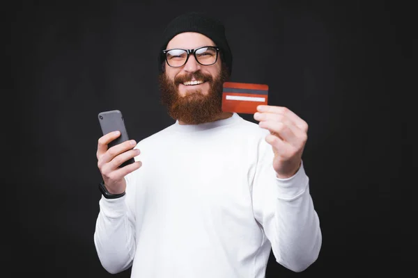 Joven alegre mostrando su nueva tarjeta de crédito y sosteniendo el teléfono inteligente — Foto de Stock