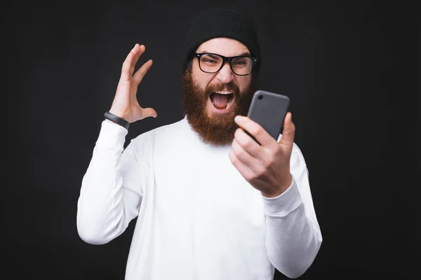 Irritado barbudo homem gritando no smartphone em pé sobre fundo escuro — Fotografia de Stock