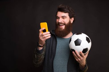 Happy man looking at smartphone and holding soccer ball, fan support team