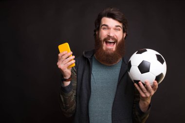 Happy man looking at smartphone and holding soccer ball, fan support team