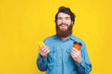 Neşeli sakallı hippi adam elinde kahve ve akıllı telefon tutuyor.