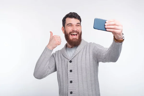Jeune homme barbu souriant prend un selfie et montrant un pouce sur le mur blanc . — Photo