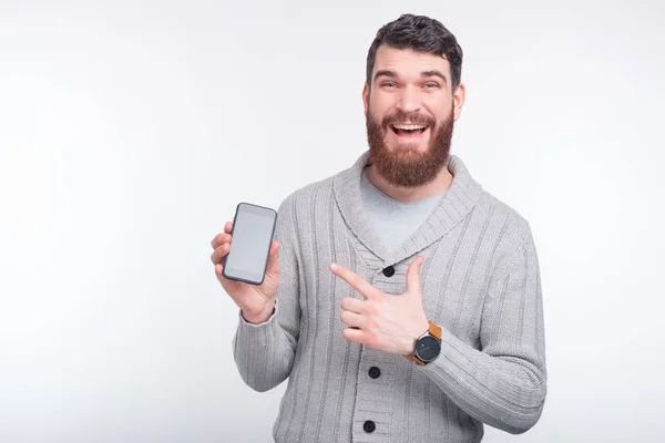 Jeune homme barbu sourit à la caméra et pointant vers smartphone près d'un mur blanc . — Photo