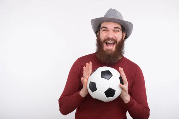 Um jovem homem barbudo feliz está segurando com ambas as mãos uma bola de futebol perto de uma parede branca . — Fotografia de Stock
