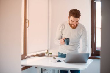 Gülümseyen adam beyaz ofisinde ya da mutfağında bardak tutarken bilgisayarını kontrol ediyor..