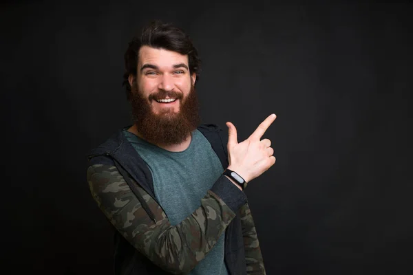 Mira a mi izquierda. Sorprendido, sonriente hombre barbudo señala a su izquierda sobre fondo negro . — Foto de Stock