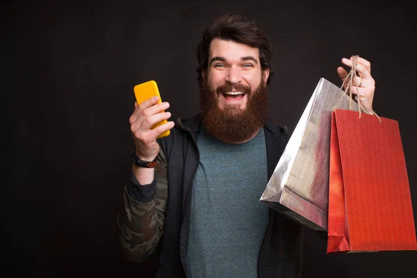Compras on-line é ótimo. Homem barbudo está segurando dois sacos de compras e seu telefone no fundo preto . — Fotografia de Stock