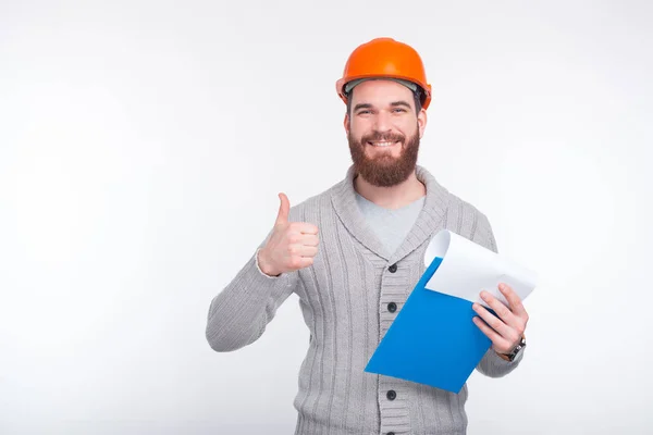 Me gusta mi trabajo. El joven ingeniero lleva puesto un sombrero naranja mientras sostiene unos papeles que muestran un gesto. —  Fotos de Stock