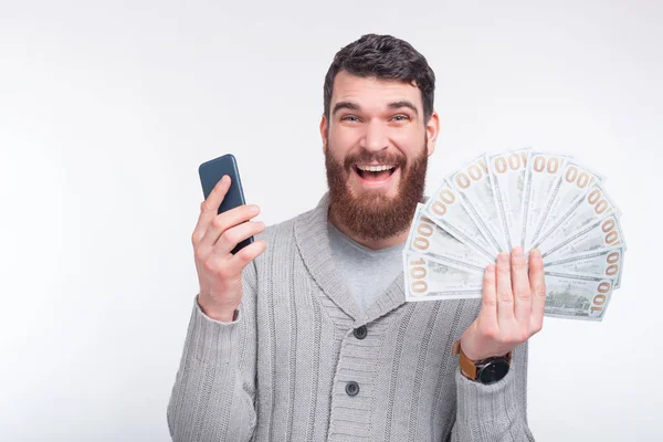 Look how much cash i have won on betting sites, online. An exciting bearded man is showing at the camera his phone and some money. — Stock Photo, Image