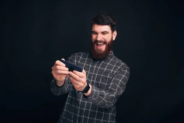 Que grande jogo. Adulto jovem barbudo homem está jogando jogos em seu telefone no fundo escuro . — Fotografia de Stock