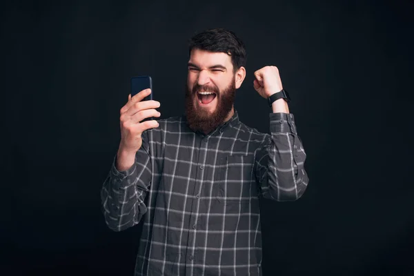 Oh, sí, he ganado. Hombre barbudo excitado está mirando su teléfono y haciendo el gesto ganador con un brazo . — Foto de Stock