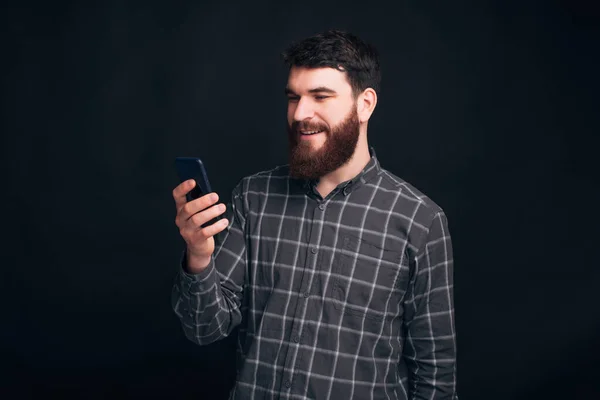 Young bearded student is searching his phone and smiling at it on black background. — Stock Photo, Image