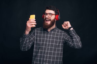 Neşeli genç sakallı adam kulaklıkla müzik dinliyor şarkı söylüyor ve telefonuna bakıyor..