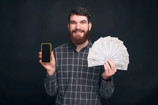 大胡子的年轻人正拿着他的手机，手里拿着一堆钱。你可以通过你的电话工作. — 图库照片