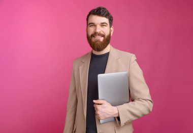 Takım elbiseli yakışıklı bir adam elinde dizüstü bilgisayarla kameraya bakıyor.