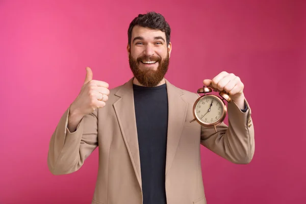 Feliz joven hombre de negocios mostrando los pulgares hacia arriba gesto y la celebración de reloj despertador sobre fondo rosa — Foto de Stock