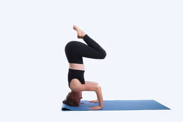 Mooie jonge vrouw uit te werken in een studio op witte achtergrond, doen yoga oefening op blauwe mat, variatie van ondersteunde headstand, garuda salamba sirsasana. — Stockfoto