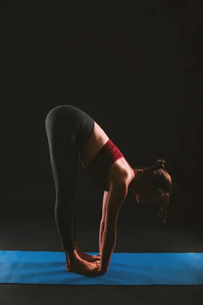 Молода приваблива спортивна жінка, що практикує йогу, стоїть вперед, вправа на коліна, утотанасана позу, працює, одягнена в спортивний одяг на чорному тлі . — стокове фото