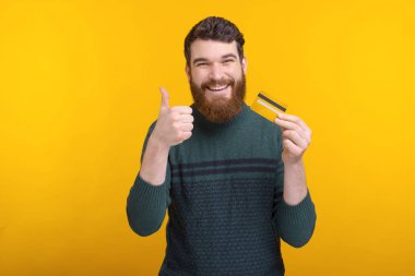 Cheerful man with beard holding credit card and showing thumbs up gesture