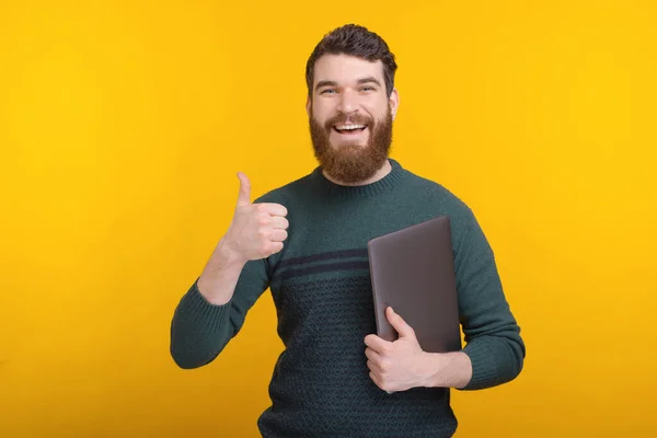 Joven alegre con barba mostrando los pulgares hacia arriba gesto y sosteniendo su computadora portátil — Foto de Stock