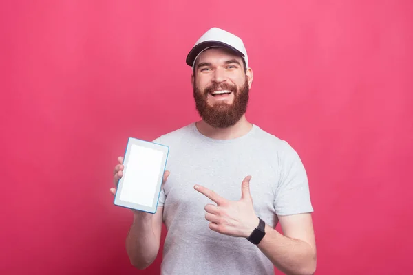 Feliz hombre sonriente con barba apuntando a la pantalla de la tableta sobre fondo rosa — Foto de Stock