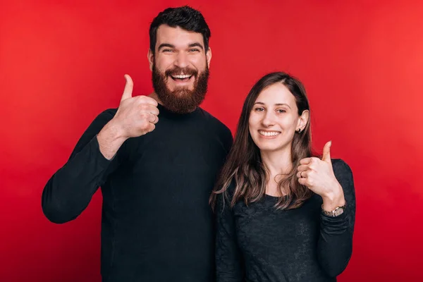 Foto de alegre pareja joven de pie sobre el fondo rojo y mostrando los pulgares hacia arriba —  Fotos de Stock