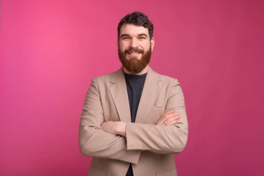 Kollarını kavuşturmuş kendine güvenen sakallı adam kameraya gülümsüyor. 