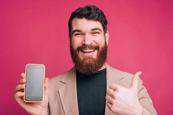 Joven barbudo alegre está mostrando su teléfono de pantalla en blanco y — Foto de Stock