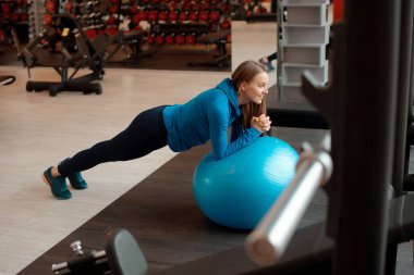 Genç bir kadın spor salonunda fitness topuyla kalas yapıyor.