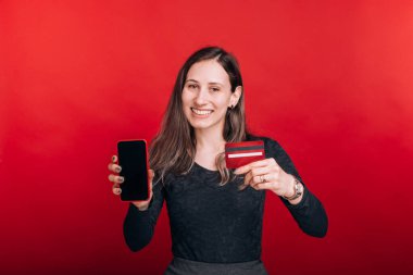 Mobil bankacılık harika bir şey. Kırmızı arka plandaki neşeli genç bayan cep telefonunu ve kredi kartını gösteriyor.
